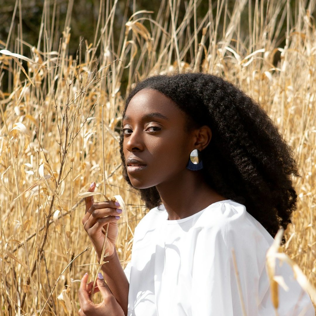 Plant Medicine, Natural Health, and Herbal Care A black woman with curly hair stands among tall golden grass, wearing a white blouse and earrings.